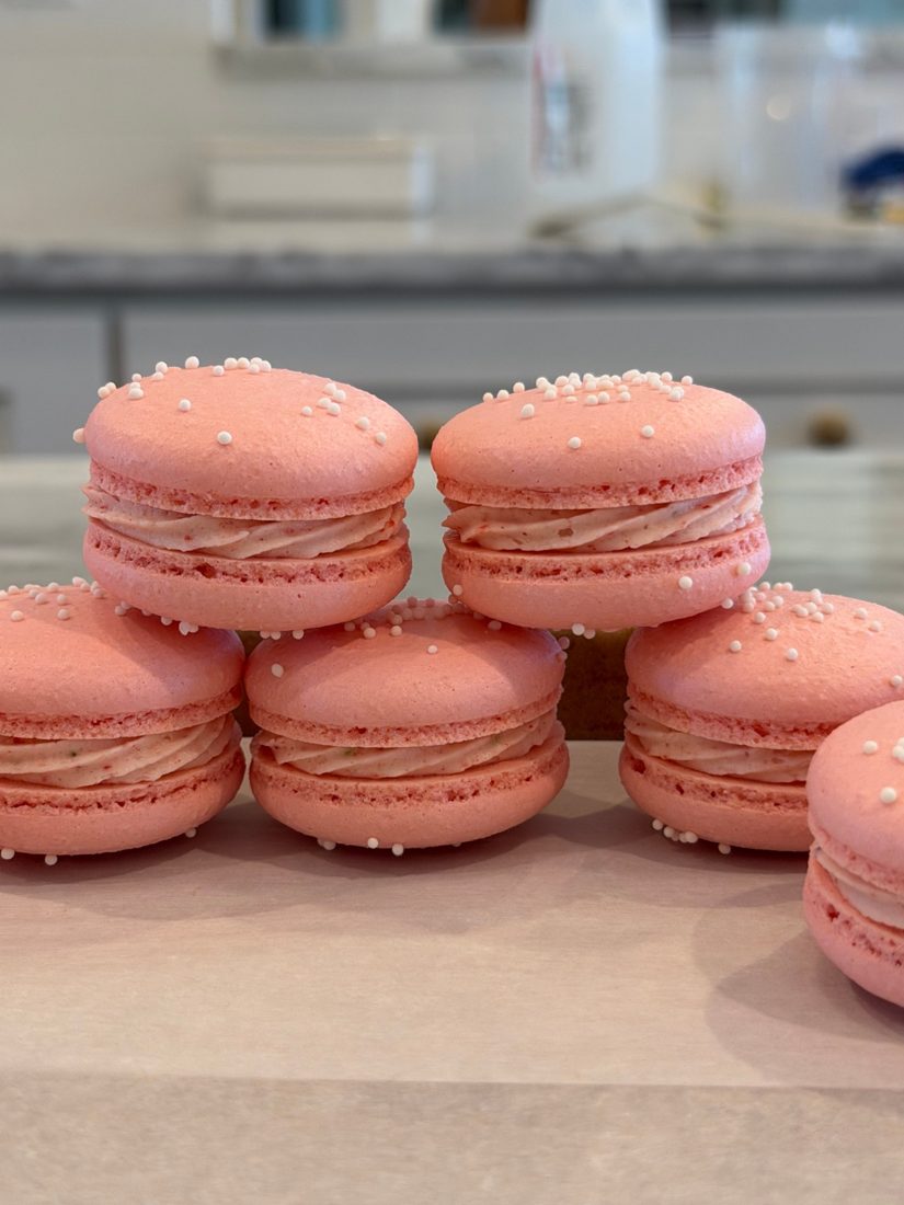 A stack of pink macarons with white sprinkles and a swirl of filling on a wooden board in a bright kitchen